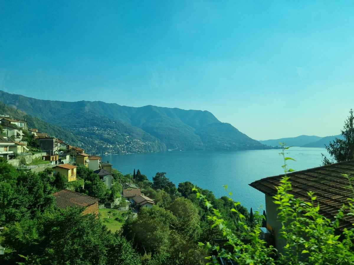 Πανοραμική φωτογραφία που δείχνει τη Λίμνη Κόμο με γραφικά ιταλικά σπίτια σε λόφο, γαλάζια νερά και βουνά στο φόντο.