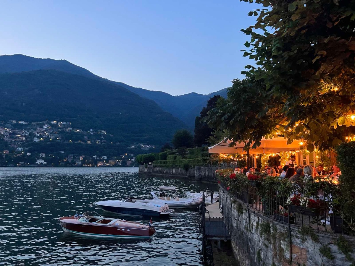 Παραλίμνιο εστιατόριο στη Λίμνη Κόμο την ώρα του aperitivo, με βαρκάκια δεμένα στο νερό και φωτισμένη βεράντα.