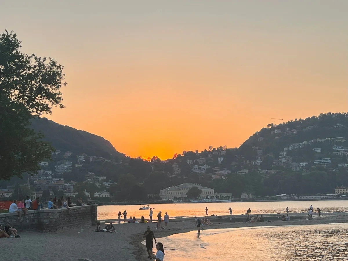 Ηλιοβασίλεμα στη Λίμνη Κόμο με κόσμο να περπατά και να χαλαρώνει στην παραλία, απολαμβάνοντας τη θέα και την ατμόσφαιρα του καλοκαιριού.