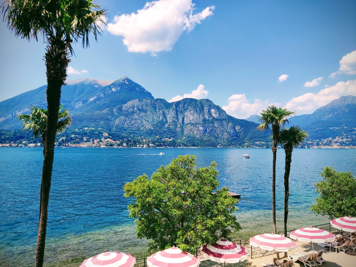 Θέα στη Λίμνη Κόμο με παραλία, φοίνικες και βουνά, που αντικατοπτρίζει την ομορφιά των κοντινών χωριών Lecco, Tremezzo και Varenna.