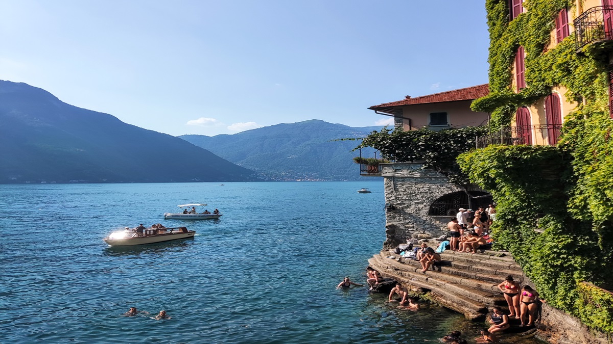 Κόσμος κολυμπά και απολαμβάνει τις βάρκες στο παραθαλάσσιο χωριό Nesso στη Λίμνη Κόμο, με βουνά στο φόντο.