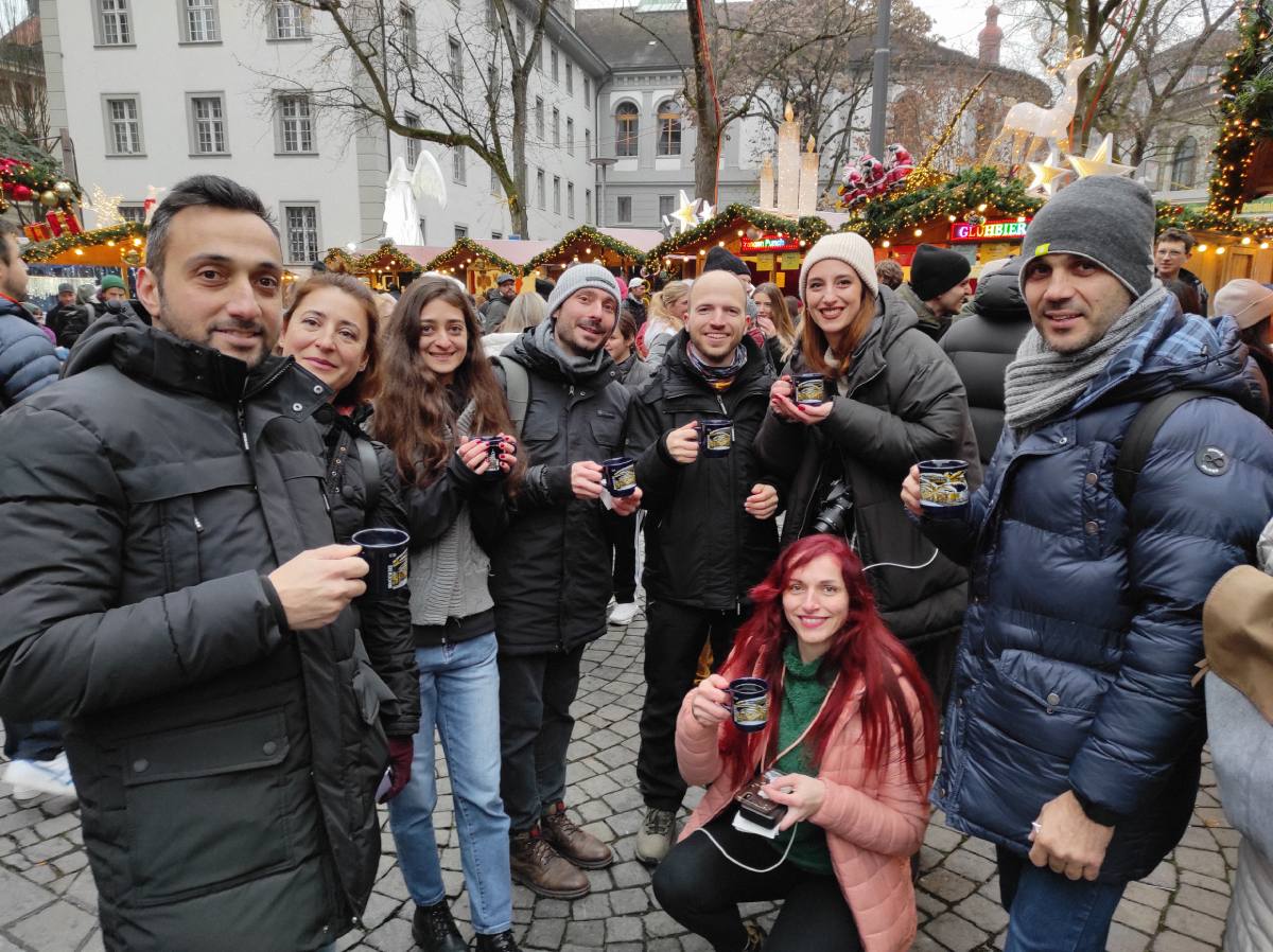 Παρέα στις χριστουγεννιάτικες αγορές του Κολμάρ με κούπες vin chaud, στολισμένα περίπτερα στο βάθος