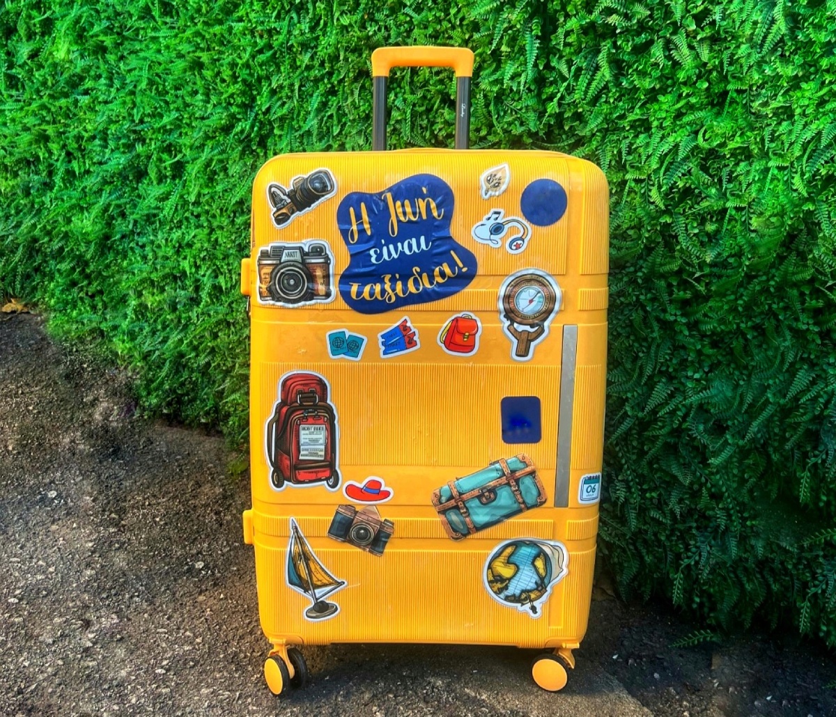 Μια κίτρινη βαλίτσα καμπίνας με πολλά πολύχρωμα αυτοκόλλητα με θέμα τα ταξίδια, όπως κάμερες, πυξίδα και μια φράση που λέει "Η ζωή είναι ταξίδια!"